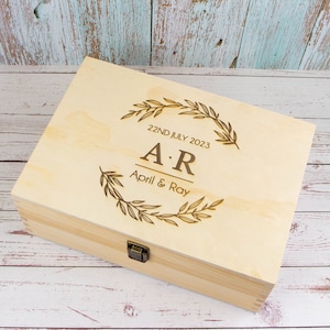 May include: A light wood keepsake box with a hinged lid and a brass latch. The lid is engraved with a wreath of leaves, the initials "A.R." and the text "22ND JULY 2023 April & Ray".