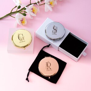 May include: Three personalized compact mirrors in gold, silver, and rose gold, each with a floral initial design and name. One is in a white box, another in a black velvet pouch, and the third is on a clear block. The background is pink with white flowers.