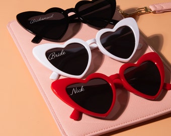 May include: Three pairs of heart-shaped sunglasses with different colored frames: black, white, and red. Each pair has a different name inscribed on the frame: "Bride", "Bridesmaid", and "Nick".