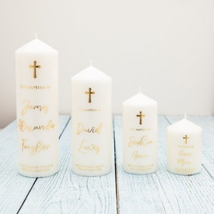 May include: Four white pillar candles with gold crosses and gold lettering. The candles are personalised with names and dates. The candles are arranged in order of size from largest to smallest.