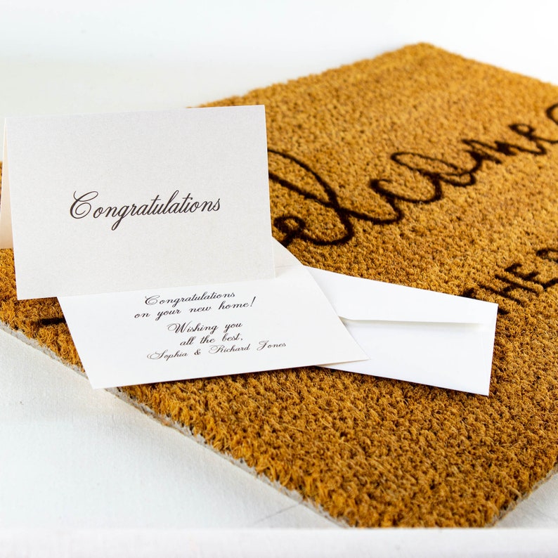 May include: Two white cards with black calligraphy on a brown welcome mat. The top card says "Congratulations". The bottom card says "Congratulations on your new home! Wishing you all the best. Sophia & Richard Jones".