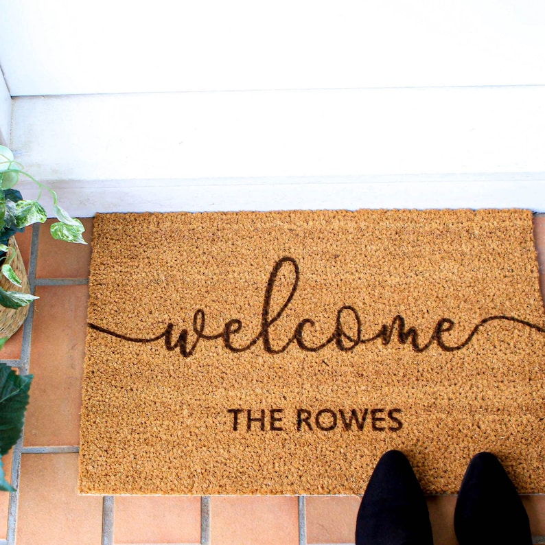 May include: A brown coir welcome mat with the text "welcome" in a cursive font and "THE ROWES" in a block font.