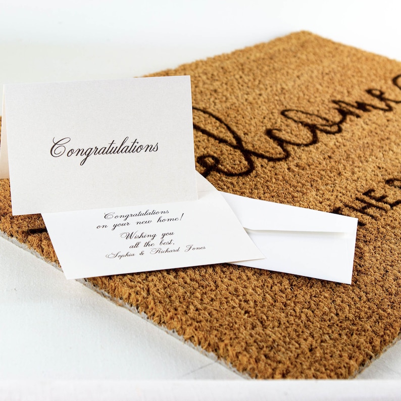 May include: A white card with the text "Congratulations" on the top and "Congratulations on your new home! Wishing you all the best. Sophia & Richard Jones" on the bottom. The card is sitting on a brown welcome mat.