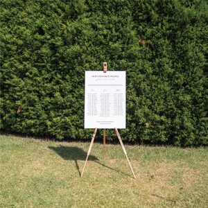 May include: A white seating chart with the text "OUR FAVORITE PEOPLE" and guest names, displayed on a wooden easel. The chart is set against a backdrop of green foliage and grass.