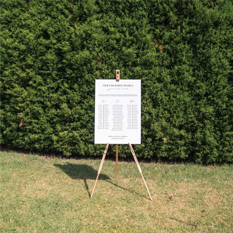 May include: A white seating chart with the text "OUR FAVORITE PEOPLE" and guest names, displayed on a wooden easel. The chart is set against a backdrop of green foliage and grass.