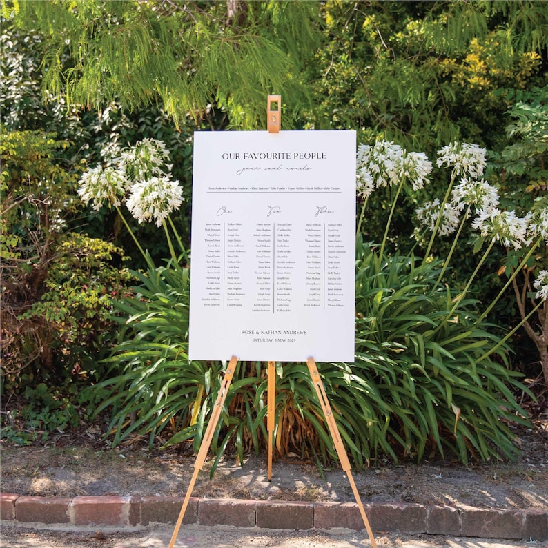 May include: A white wedding seating chart on a wooden easel, with the text "OUR FAVOURITE PEOPLE" at the top. The names are arranged in columns under signatures. The sign is set outdoors with greenery in the background.