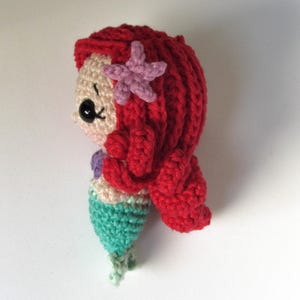 May include: A small crochet mermaid doll featuring bright red yarn hair, a light purple starfish hair accessory, a beige face with black plastic safety eyes, a purple seashell top, and a textured mint green tail with a light green fin at the base.