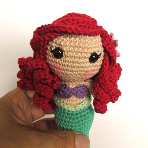 Op de afbeelding: Handgemaakte gehaakte pop van een zeemeermin met rood haar, een paarse top en een groene staart. De pop heeft grote zwarte ogen en een kleine roze bloem in haar haar. De pop wordt in een hand gehouden.