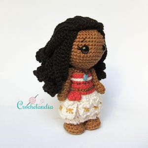 May include: Handmade crochet doll depicting a dark-skinned character with black hair. The doll is dressed in a red and white top, white trousers with gold details, and a blue pendant. The doll has a cartoonish design.