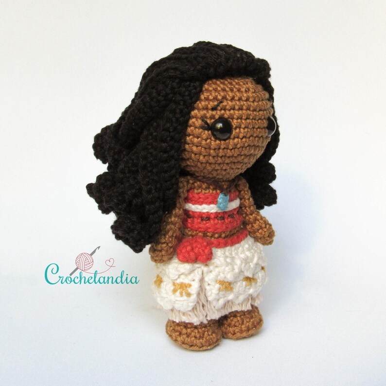 May include: Handmade crochet doll depicting a dark-skinned character with black hair. The doll is dressed in a red and white top, white trousers with gold details, and a blue pendant. The doll has a cartoonish design.