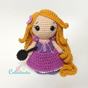 May include: Crocheted doll with long blonde hair, wearing a purple dress and holding a black frying pan. The doll has a pink flower in her hair and brown eyes.
