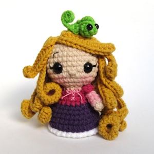May include: A crocheted doll with long golden yarn hair, featuring a small green chameleon perched on top. The doll wears a pink and purple dress with white trim at the base, black bead eyes, and pink blush on the cheeks, crafted in an amigurumi style.