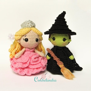 May include: A crocheted doll dressed as a princess in a pink dress and a silver crown, standing next to a crocheted doll dressed as a witch in a black dress and a black pointed hat, holding a brown broom.