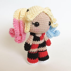 May include: A handmade crochet doll with a light skin tone, wearing a red, black, and white diamond-patterned outfit. The doll has blonde hair with pink and blue accents, and large black eyes. The doll is approximately 6 inches tall.