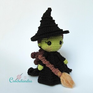 May include: A handmade crochet witch doll with a black hat and dress, green skin, and a brown broom. The doll has large black eyes and is holding a brown broom. The doll is a whimsical, handcrafted item.