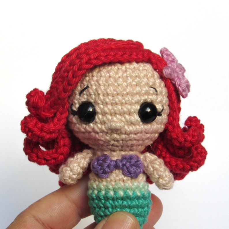 May include: A small amigurumi crochet doll featuring a character with vibrant red yarn hair, a light beige face, large black plastic eyes, a purple crocheted seashell top, and a teal green crocheted mermaid tail. A small light purple flower is attached.