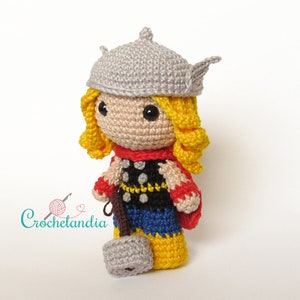 May include: Crocheted plush toy of a superhero with a grey helmet, yellow hair, red cape, and a black and yellow costume holding a grey hammer.
