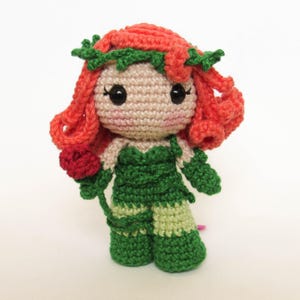 May include: A handmade crochet doll with red hair, green clothing, and a red rose. The doll has large black eyes and a light skin tone. The doll's hair is styled with a green vine crown. The doll's outfit is green with lighter green accents.
