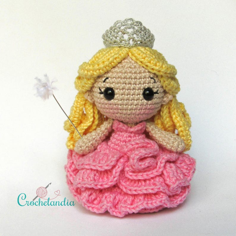 May include: A handmade crochet doll in the shape of a princess. The doll has yellow hair, a silver crown, and a pink dress. She is holding a white wand. The doll is made with yarn and has black eyes. The logo "Crochetlandia" is in the bottom left corner.