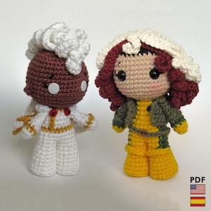 Puede incluir: Dos muñecas de ganchillo hechas a mano. Una muñeca viste un mono blanco con detalles dorados y un peinado blanco y esponjoso. La otra muñeca lleva un conjunto amarillo con una chaqueta verde y pelo rojo con una mecha blanca. Las muñecas tienen ojos grandes y negros.
