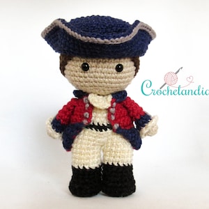 Può includere: Una bambola all'uncinetto che indossa un cappello blu e grigio, una giacca rossa e blu e pantaloni bianchi. La bambola ha occhi neri e un viso marrone.