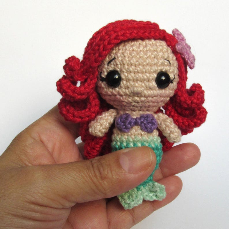 May include: A small crocheted mermaid doll featuring bright red yarn hair, a light beige face with large black safety eyes, a purple shell-shaped top, and a light green tail. A small pink flower is attached to the side of the red hair.