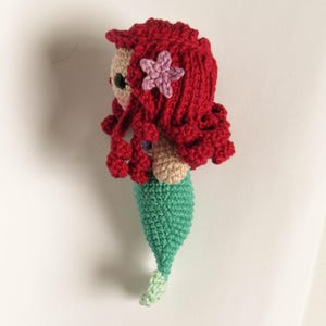 Op de afbeelding: Handgemaakte gehaakte zeemeerminnenpop met rood haar, een roze zeester haaraccessoire en een groene staart. De pop heeft een bruin gezicht en is gemaakt van garen.