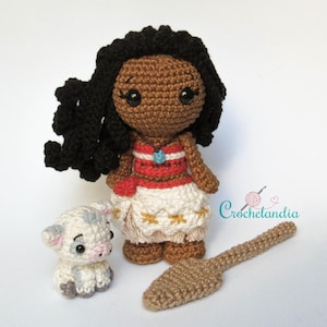 May include: A handmade crochet doll with dark skin, black hair, and a red and white dress. A small crocheted pig and a brown crocheted paddle are also present. The doll has a blue pendant. The word "Crochetlandia" is in the image.