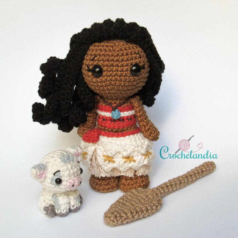 May include: A handmade crochet doll with dark skin, black hair, and a red and white dress. A small crocheted pig and a brown crocheted paddle are also present. The doll has a blue pendant. The word "Crochetlandia" is in the image.