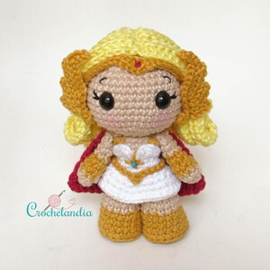 May include: A handmade crochet doll of a character with blonde hair, a red cape, and a white dress with gold accents. The doll has large black eyes, rosy cheeks, and is wearing gold boots. The doll is approximately 6 inches tall.