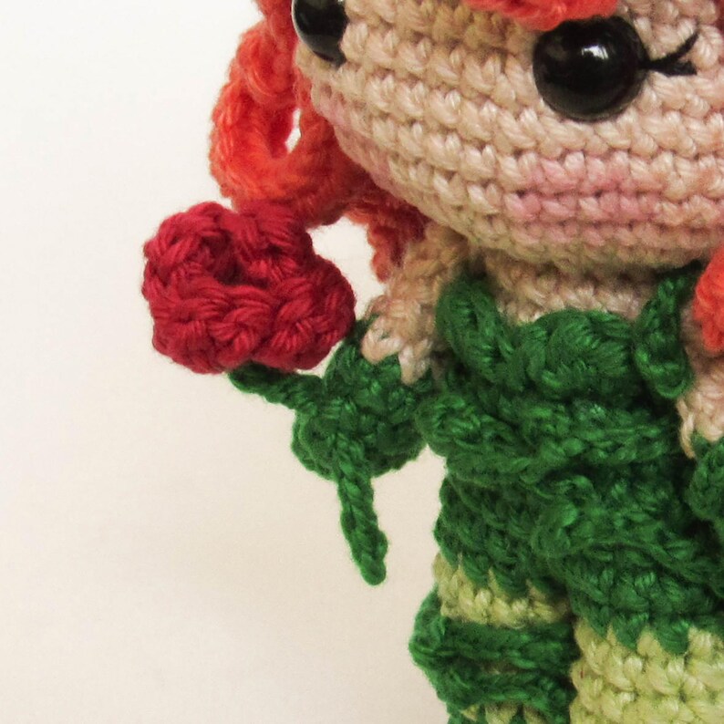 May include: Crocheted doll with red hair, black eyes, and a green and beige outfit. The doll is holding a red flower. The crochet work is detailed, with visible stitches and textured surfaces. The doll's features are carefully crafted.