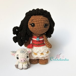 May include: Crocheted doll of a character with dark skin, black curly hair, and a red and white dress. A small crocheted pig is next to the doll. The doll has a blue necklace. The doll is approximately 18 cm tall.