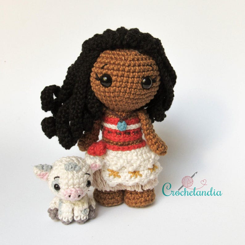 May include: Crocheted doll of a character with dark skin, black curly hair, and a red and white dress. A small crocheted pig is next to the doll. The doll has a blue necklace. The doll is approximately 18 cm tall.