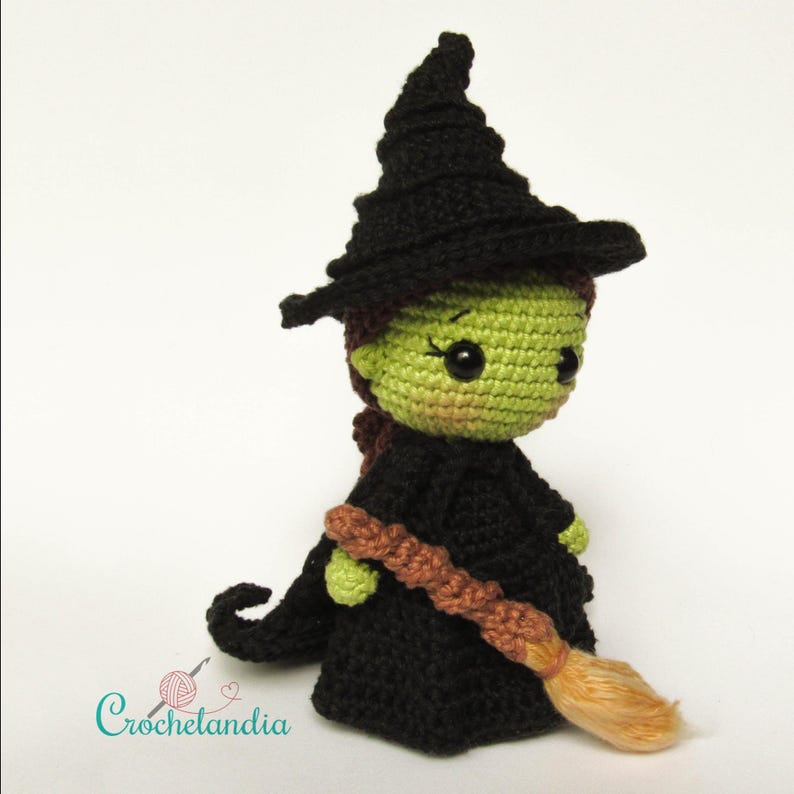 May include: A handmade crochet witch doll with a green face, black hat and dress, and a brown broom. The doll has black eyes and a small, pointed nose. The word "Crochetlandia" is in the lower left corner.