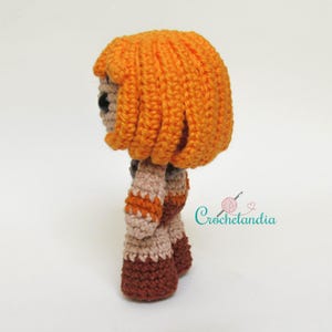 May include: A handmade crochet doll with orange hair, black eyes, and a tan face. The doll is wearing a tan and brown outfit. The doll's hair is styled in a short, layered bob. The doll is a side view. The text "Crochetlandia" is in the image.