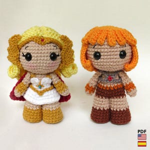 Puede incluir: Dos muñecas de ganchillo hechas a mano. Una muñeca tiene cabello amarillo, una capa roja y un vestido blanco. La otra muñeca tiene cabello naranja y un atuendo marrón y naranja. Ambas muñecas tienen ojos negros grandes y están de pie.