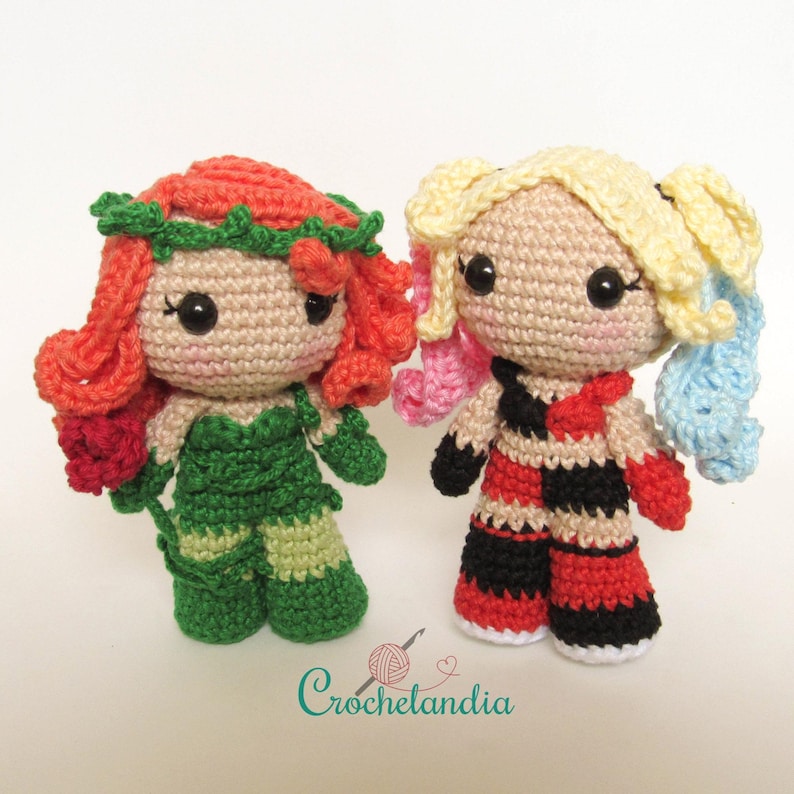 May include: Two handmade crochet dolls, one resembling Poison Ivy with red hair and a green outfit, and the other Harley Quinn with blonde pigtails and a red, black, and beige outfit. The dolls have large black eyes and are standing side-by-side.