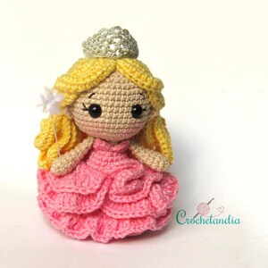 May include: A handmade crochet doll in the shape of a princess. The doll has blonde hair, a silver crown, and a pink dress. She is holding a star-shaped wand. The doll is a charming handmade item.