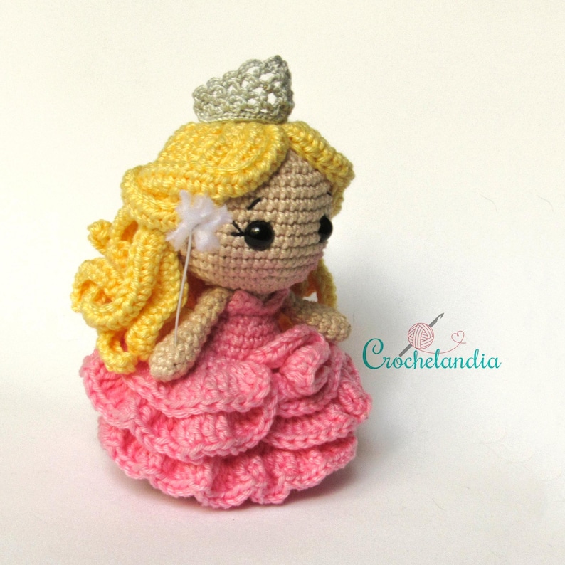May include: A handmade crochet doll of a princess with yellow hair, a pink dress, and a silver crown. The doll has black eyes and a small white wand. The doll is a whimsical, handcrafted item.