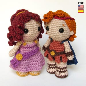 May include: Two handmade crochet dolls, one in a purple dress with a gold sash and the other in a brown and red outfit with a blue cape. Both dolls have black eyes and are made with yarn.