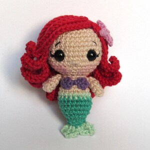 May include: A small crocheted mermaid doll featuring bright red yarn hair with a light purple flower accessory, a beige yarn face with black plastic eyes, a purple yarn seashell top, and a green yarn tail with a light green fin at the base.