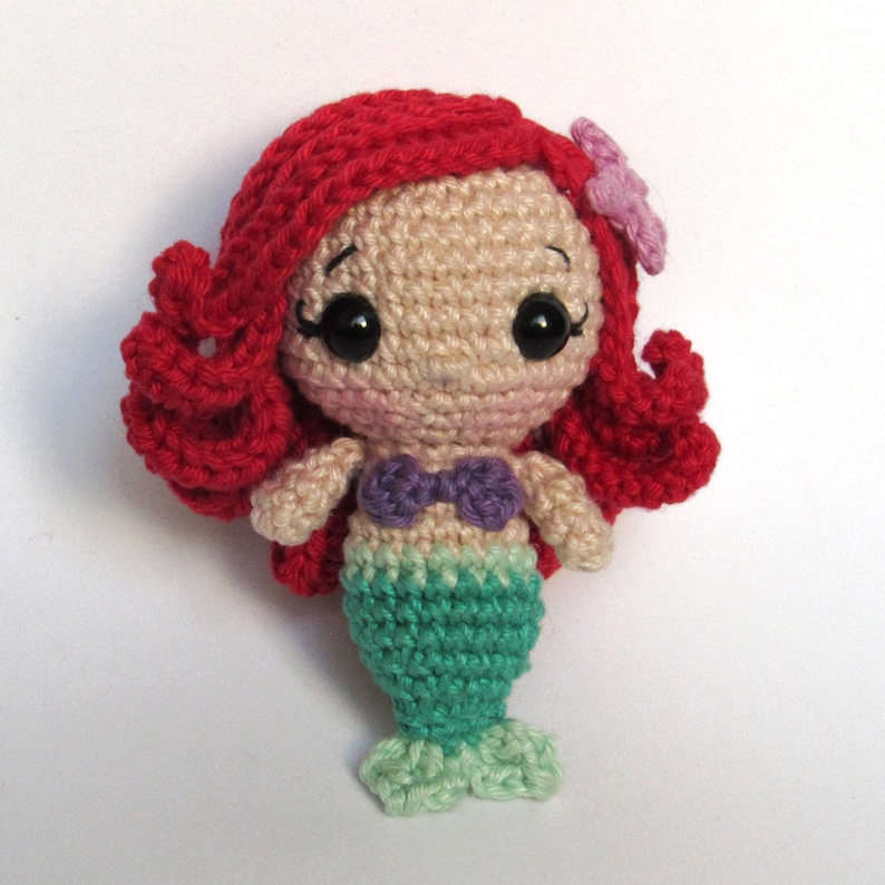 May include: A small crocheted mermaid doll featuring bright red yarn hair with a light purple flower accessory, a beige yarn face with black plastic eyes, a purple yarn seashell top, and a green yarn tail with a light green fin at the base.