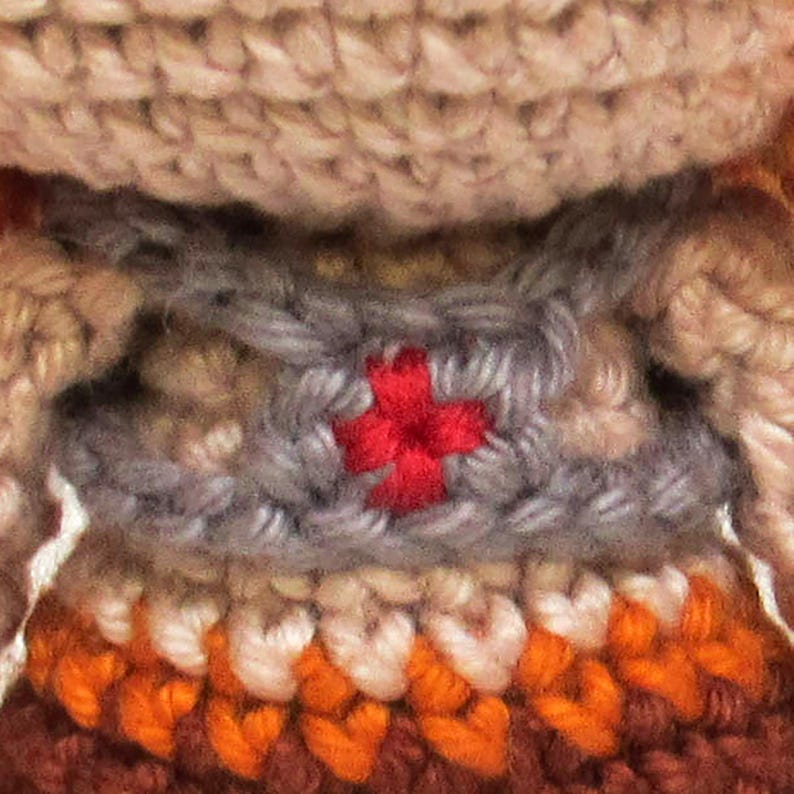 May include: Close-up of a handmade crochet item. The item features a red cross stitched onto a gray section, with tan, orange, and brown sections below. The crochet work is detailed and textured, showcasing the craftsmanship.
