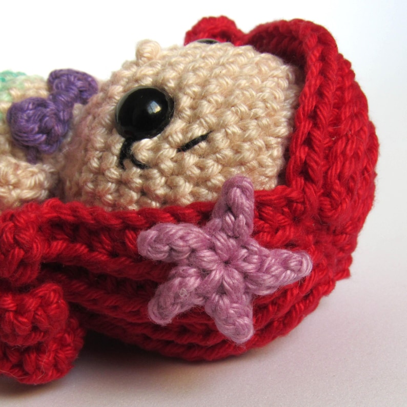 May include: A beige crocheted animal figure with a black plastic nose and a small purple bow, nestled inside a bright red crocheted cradle. The cradle is decorated with a light purple crocheted starfish accent on the side, featuring textured yarn.