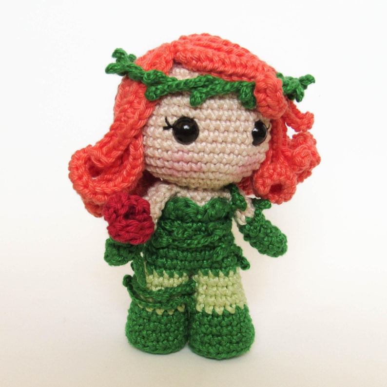 May include: Crocheted doll featuring a character with red hair, green leafy accents, and a green and light green outfit. The doll has large black eyes and a red rose in its hand. The doll is standing on a white surface.