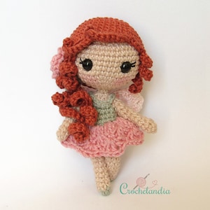 Pode incluir: Uma boneca de fada de crochê com um vestido rosa, um corpete verde e cabelo comprido e encaracolado vermelho. A boneca tem uma flor no cabelo e está usando um par de sapatos verdes.