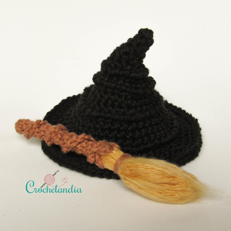 May include: A black crocheted witch hat with a wide brim and a pointed top. A small, brown and tan crocheted broom is resting on the brim. The image includes the text "Crochetlandia" in a teal script.