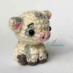 May include: A crocheted piglet in cream, grey, pink, and black. The piglet features black safety eyes, grey spots, pink ears, and a pink snout. The crochet piglet is in a sitting position. The text "Crochetlandia" is visible in the background.