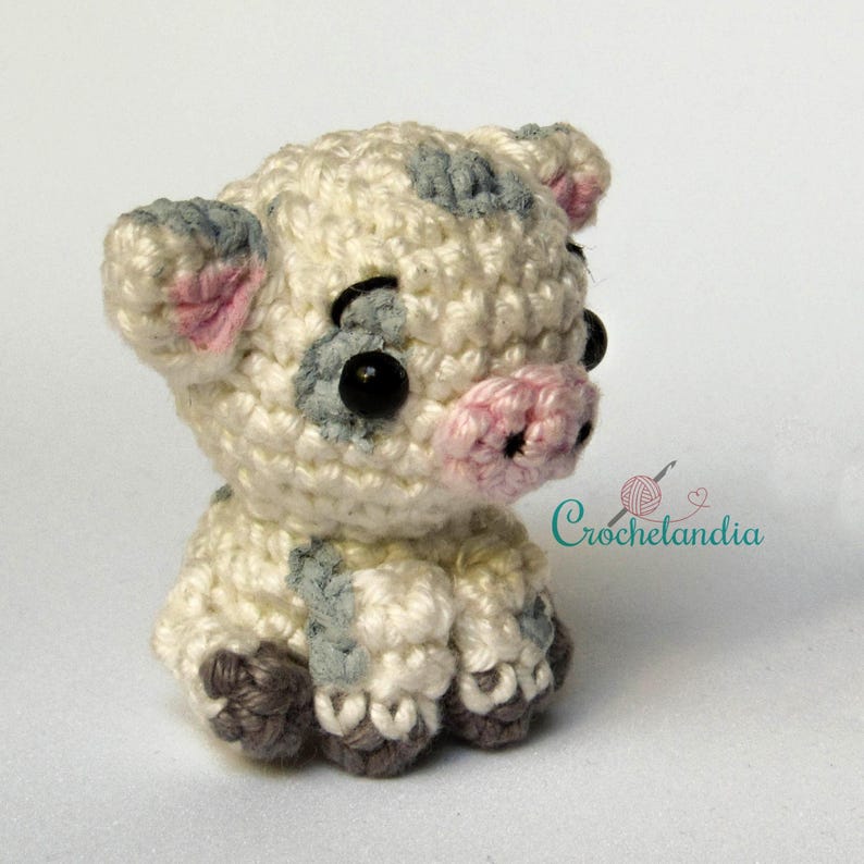May include: A crocheted piglet in cream, grey, pink, and black. The piglet features black safety eyes, grey spots, pink ears, and a pink snout. The crochet piglet is in a sitting position. The text "Crochetlandia" is visible in the background.