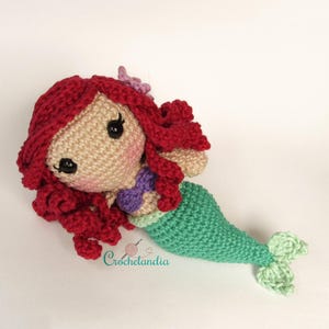 Op de afbeelding: Handgemaakte gehaakte pop van een zeemeermin met rood haar, een beige gezicht en een groene staart. De pop heeft zwarte ogen, roze wangen en een paarse strik. Een grillig, handgemaakt item.
